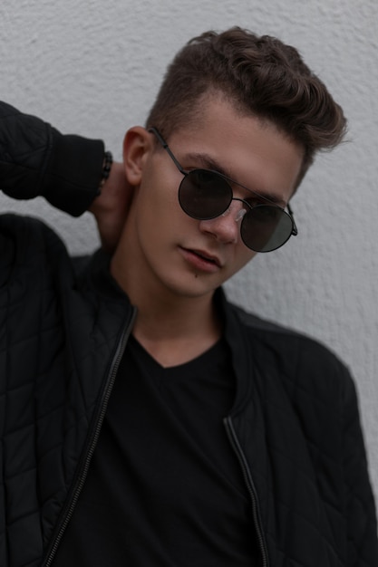 Ritratto alla moda di un bel ragazzo giovane con occhiali da sole alla moda in vestiti neri vicino al muro grigio