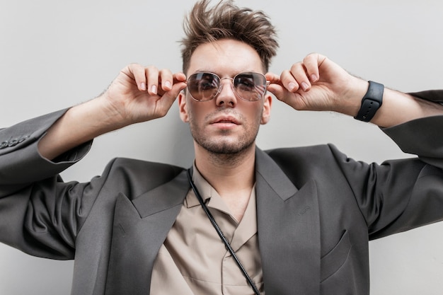 ritratto alla moda di un bell'uomo modello con barba corta in abito casual alla moda con un blazer e una camicia guarda attraverso gli occhiali da sole vicino a un muro grigio