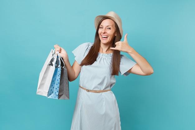 Ritratto alla moda sorridente bella donna caucasica in abito estivo, cappello di paglia che tiene pacchetti borse con acquisti dopo lo shopping isolato su sfondo blu pastello. Copia spazio per la pubblicità.