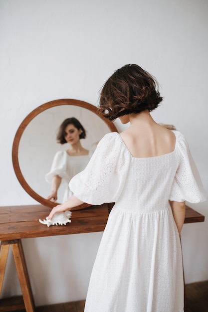 Ritratto allo specchio di una donna timida sensuale in un abito bianco. Bella bruna con i capelli corti al chiuso. Da dietro.