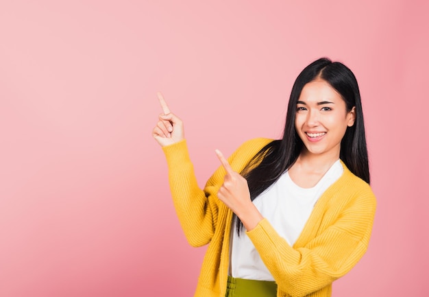Ritratto asiatico felice bella giovane donna carina in piedi fa gesto due dita puntate verso l'alto sopra presentando qualcosa di prodotto, girato in studio isolato su sfondo rosa con spazio di copia