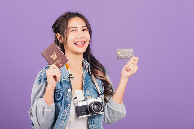 Ritratto asiatico felice bella giovane donna sorriso eccitato in estate con passaporto, carta di credito e macchina fotografica d'epoca, viaggio di viaggio pronto femminile tailandese guardando di lato lontano isolato su sfondo viola