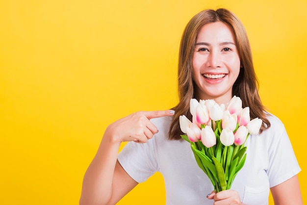 Ritratto asiatico tailandese bella giovane donna felice sorridente, urlando eccitato tenere bouquet di fiori di tulipani nelle mani e puntare il dito verso i fiori, girato in studio isolato su sfondo giallo, con spazio di copia