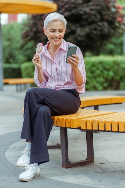 Ritratto autentico di una donna d'affari senior sorridente che tiene il gelato e usa il telefono cellulare in strada urbana stile di vita positivo Affari di successo