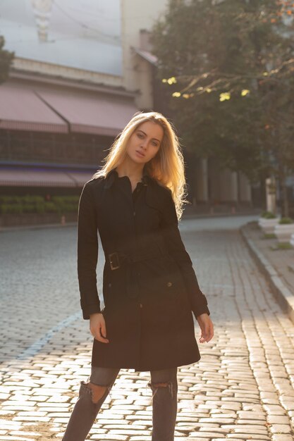 Ritratto autunnale soleggiato di una splendida modella bionda che indossa un cappotto nero e posa per la strada