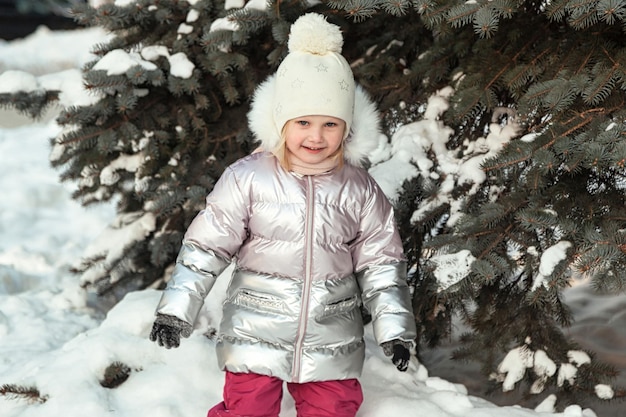 Ritratto bambina felice nel parco invernale pubblico innevato con abeti o abete rosso Bambino carino in abiti invernali luminosi
