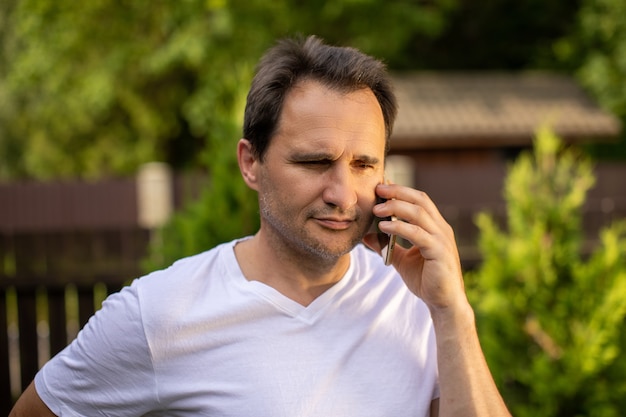 Ritratto calmo dell'uomo d'affari europeo maturo con la barba lunga positivo degli anni '40 in maglietta bianca che parla sul telefono cellulare all'aperto sulla natura verde vaga. Il concetto di business per il controllo dei sentimenti