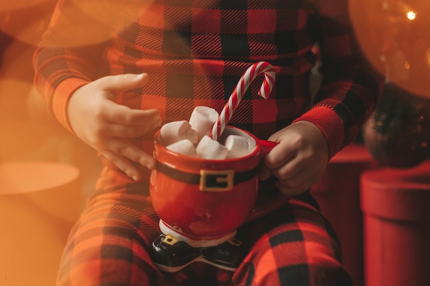 Ritratto candido bambino felice in pigiama plaid rosso tenere tazza di Natale con marshmallow e bastoncino di zucchero