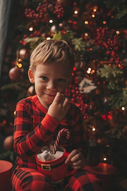 Ritratto candido bambino felice in pigiama plaid rosso tenere tazza di Natale con marshmallow e bastoncino di zucchero