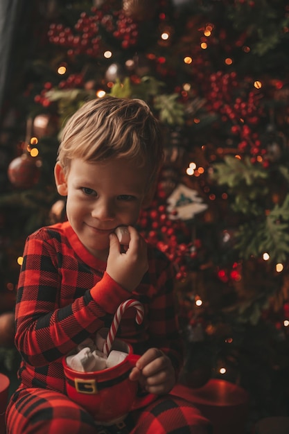 Ritratto candido bambino felice in pigiama plaid rosso tenere tazza di Natale con marshmallow e bastoncino di zucchero