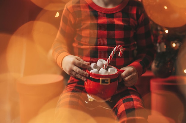 Ritratto candido bambino felice in pigiama plaid rosso tenere tazza di Natale con marshmallow e bastoncino di zucchero