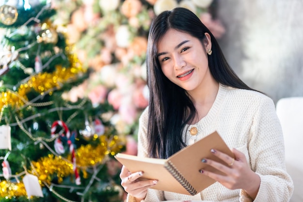 Ritratto carino bello sorriso positivo giovane donna asiatica che tiene di leggere un libro a casa nel soggiorno al chiuso Decorazione durante le vacanze di Natale e Capodanno