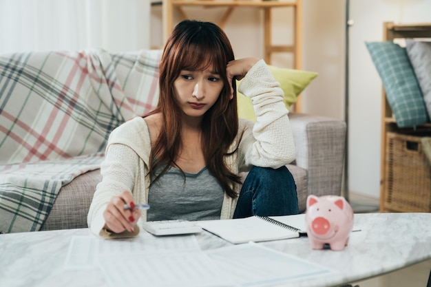 ritratto casalinga asiatica sta appoggiando la testa con un'espressione preoccupata mentre pensa a debiti e prestiti a tavola con salvadanaio nel soggiorno di casa