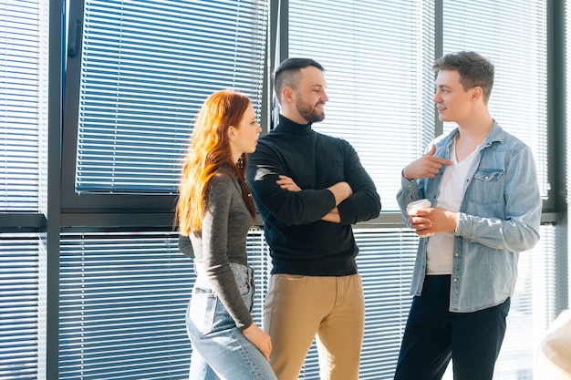Ritratto centrale di tre giovani uomini d'affari felici che si divertono conversando durante la pausa caffè in un ufficio moderno. I colleghi fiduciosi godono di una comunicazione piacevole, discutono progetto per finestra.