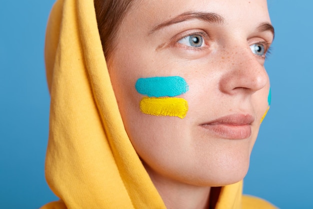 Ritratto chiuso di donna caucasica in felpa con cappuccio gialla con bandiera ucraina sulla guancia in posa isolata su sfondo blu che distoglie lo sguardo femminile patriottico con espressione facciale sognante