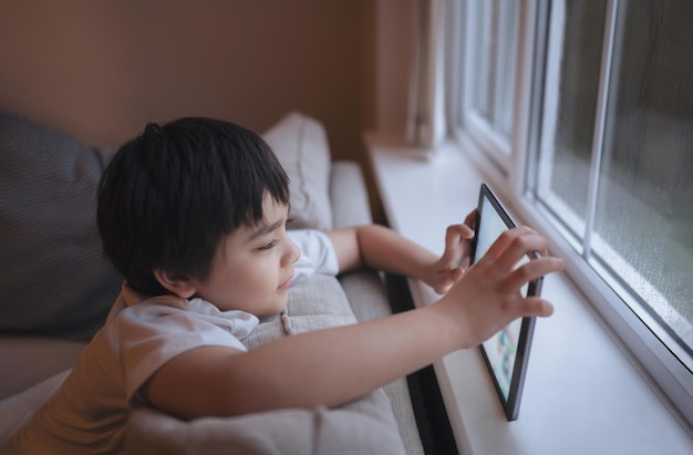 Ritratto cinematografico di un ragazzo felice che gioca online sul tablet Bambino che guarda cartoni animati da internet Bambino seduto sul divano accanto alla finestra mentre piove al mattino