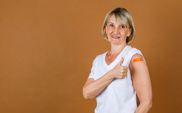 Ritratto Closeup studio shot di una paziente bionda anziana caucasica che guarda la telecamera mostra il pollice in alto sulla benda di gesso arancione sulla spalla dopo aver ricevuto la vaccinazione contro il Coronavirus.