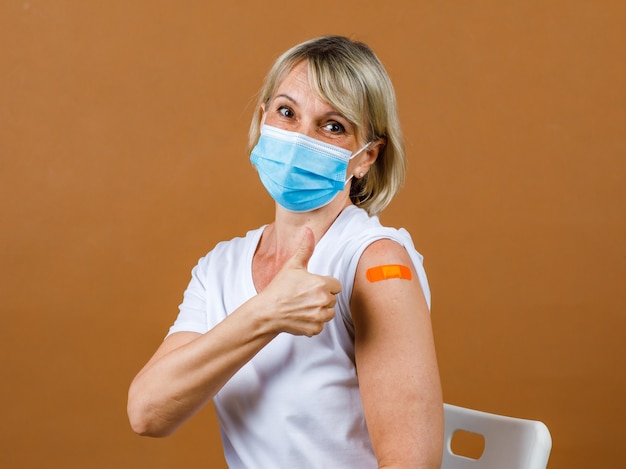 Ritratto Closeup studio shot di una paziente bionda anziana caucasica indossa una maschera facciale e guarda la telecamera mostra il pollice in alto sulla benda di gesso arancione sulla spalla dopo aver ricevuto la vaccinazione contro il Coronavirus.