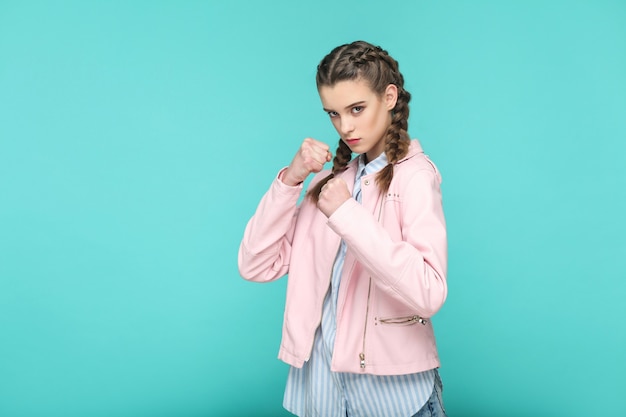 Ritratto d'attacco serio di bella ragazza carina in piedi con trucco e acconciatura a codino marrone in giacca rosa camicia a righe azzurre. indoor, studio shot isolato su sfondo blu o verde.