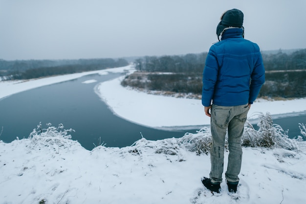 Ritratto da dietro di un uomo in abiti invernali in piedi sul bordo di una collina e guardando inverno fiume nevoso.