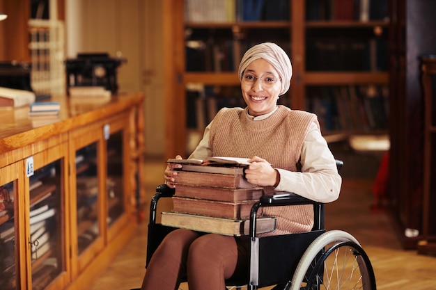 Ritratto dai toni caldi di una giovane donna sorridente che usa la sedia a rotelle nella biblioteca del college e guarda la fotocamera