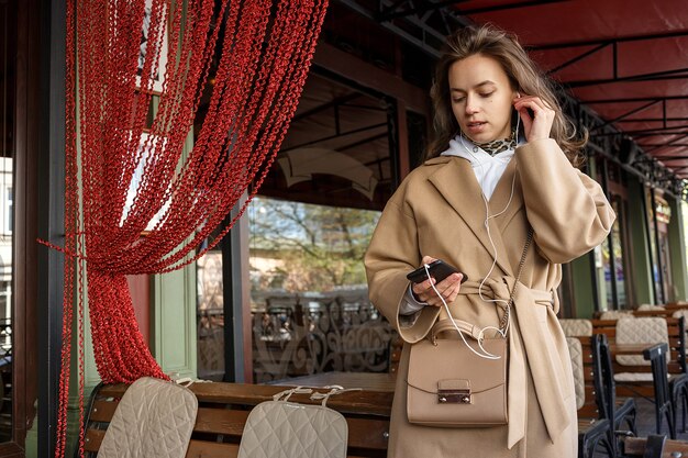 Ritratto del cappotto da portare della ragazza che ascolta la musica sulla veranda del caffè con le cuffie che tengono il telefono delle cellule in mani
