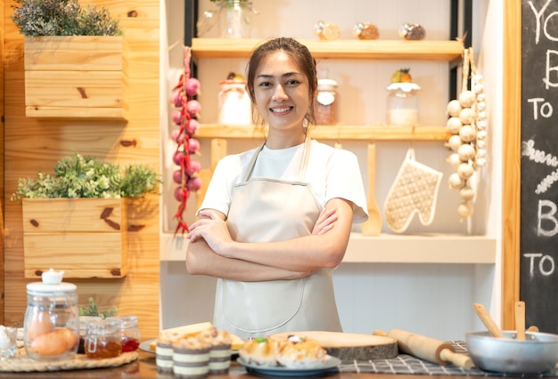 Ritratto del cuoco unico sorridente della donna asiatica di bellezza professionale che si diverte a cucinare con la pasta per il biscotto fatto in casa e l'ingrediente della torta sul tavolo in cucina