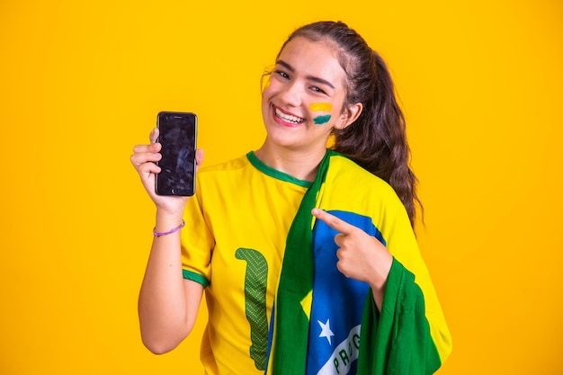 Ritratto del fan brasiliano Fan brasiliano che mostra il suo telefono cellulare vestito come un tifoso di calcio o una partita di calcio su sfondo giallo Colori della Coppa del Mondo del Brasile