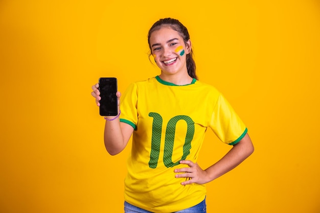 Ritratto del fan brasiliano Fan brasiliano che mostra il suo telefono cellulare vestito come un tifoso di calcio o una partita di calcio su sfondo giallo Colori della Coppa del Mondo del Brasile