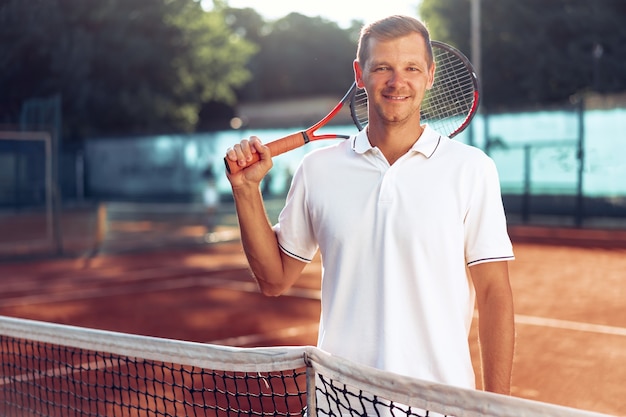 Ritratto del giocatore di tennis maschio positivo con la racchetta che sta al campo in terra battuta vicino alla rete