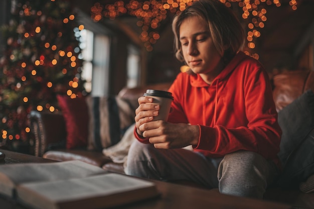 Ritratto del libro di lettura sorridente autentico schietto dell'adolescente del ragazzo e beve il caffè caldo a casa Natale