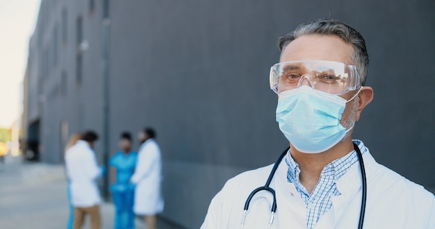 Ritratto del medico caucasico dell'uomo dai capelli grigi in mascherina medica, occhiali protettivi e con lo stetoscopio che guarda l'obbiettivo. Primo piano medico maschio in protezione respiratoria. Medici multietnici sullo sfondo.