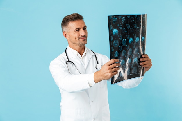 Ritratto del medico di mezza età con lo stetoscopio che tiene l'immagine dei raggi x, in piedi isolato sopra la parete blu