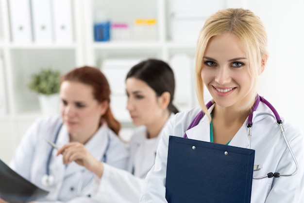 Ritratto del medico femminile sorridente della medicina che tiene la cartella blu del documento con due colleghi che esaminano l'immagine dei raggi x. Concetto di sanità e medicina.