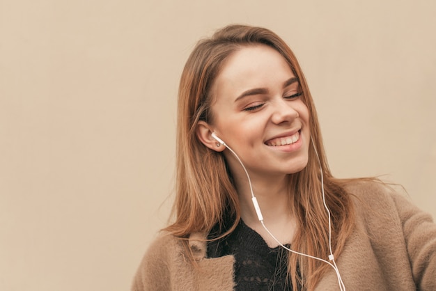 Ritratto del primo piano che ascolta la musica in sue cuffie con i suoi occhi chiusi e sorridenti, fondo beige della parete