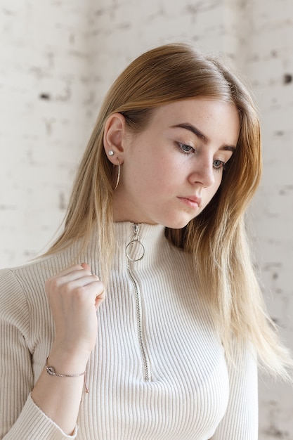 Ritratto del primo piano del modello teenager premuroso sveglio che porta maglione bianco. femmina giovane magra caucasica con capelli biondi davanti al muro di mattoni bianchi. bella signora naturale sta al loft