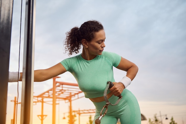 Ritratto del primo piano dell'atleta femminile che si esercita sulla macchina da palestra crossover, flettendo i muscoli della schiena, delle spalle e del tricipite con attrezzatura crossover cavo all'alba all'aperto. Concetto di fitness e bodybuilding