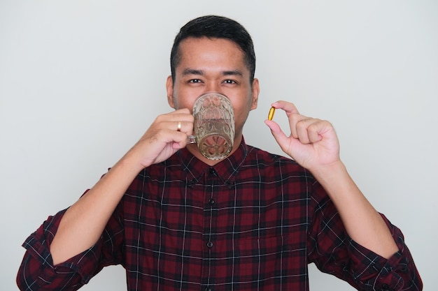 Ritratto del primo piano dell'uomo asiatico adulto che beve un bicchiere d'acqua e che tiene capsul del supplemento dell'olio di pesce