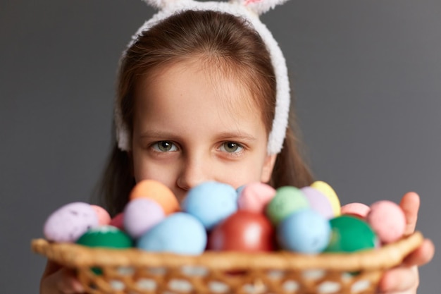 Ritratto del primo piano della bambina divertente che si nasconde dietro le uova di Pasqua nel cesto di vimini e che guarda l'obbiettivo con il viso carino che celebra la vacanza isolata su sfondo grigio