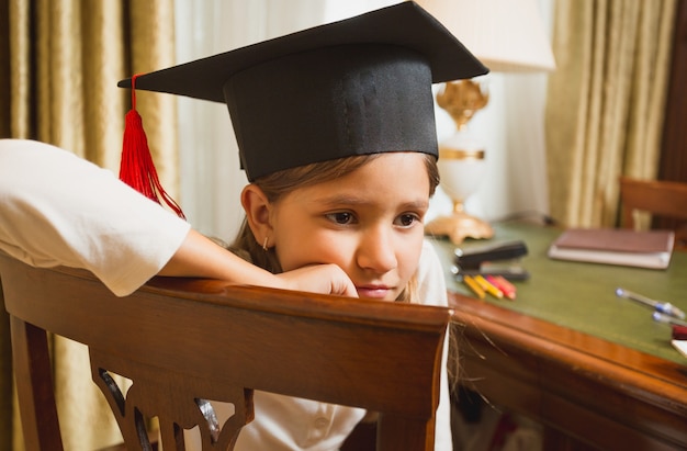 Ritratto del primo piano della bambina premurosa in cappello di graduazione che posa sulla sedia