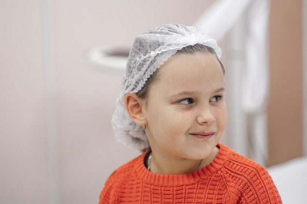 Ritratto del primo piano della bambina sorridente in maglione rosso e cappuccio medico usa e getta sulla testa in attesa di visita medica o trattamento in ospedale o clinica