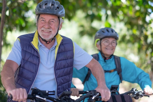 Ritratto del primo piano della coppia senior caucasica attiva con le biciclette elettriche che corrono all'aperto nel parco Due anziani sorridenti che indossano il casco da ciclismo che si godono uno stile di vita sano nella natura