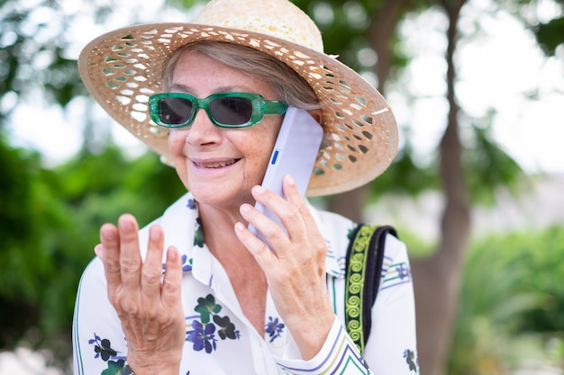 Ritratto del primo piano della donna anziana sorridente attraente con cappello e occhiali da sole che parla sul telefono cellulare nel parco pubblico signora caucasica che tiene smartphone gesticolando con la mano