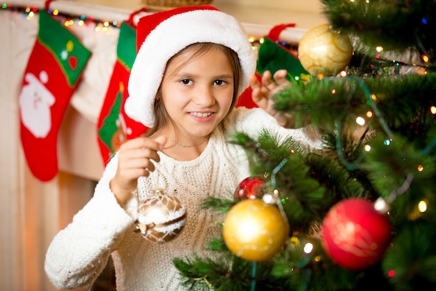 Ritratto del primo piano della ragazza sorridente sveglia che decora l'albero di Natale con le palle dorate
