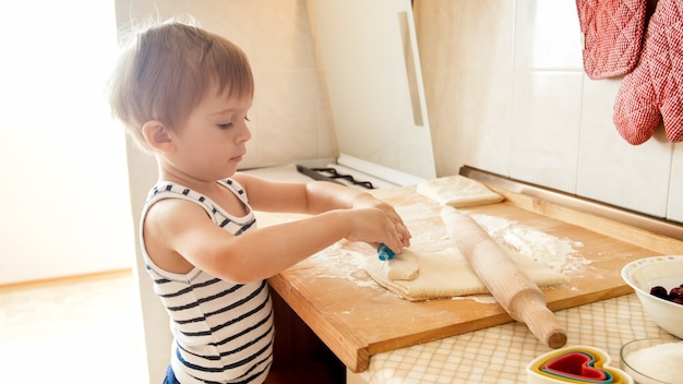 Ritratto del primo piano di adorabile bambino di 3 anni che rotola la pasta di grano con il mattarello e taglia i biscotti con una taglierina di plastica speciale. Chuld cottura e cottura in cucina