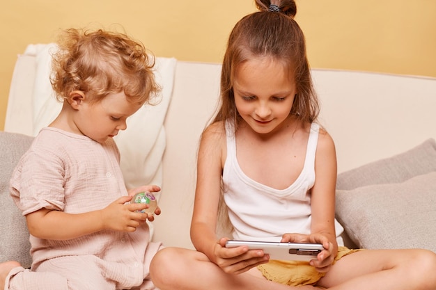 Ritratto del primo piano di affascinanti sorelle bambine curiose che utilizzano il telefono mentre si siedono sul divano di casa contro il muro beige utilizzando lo smartphone che naviga in contenuti per bambini su Internet