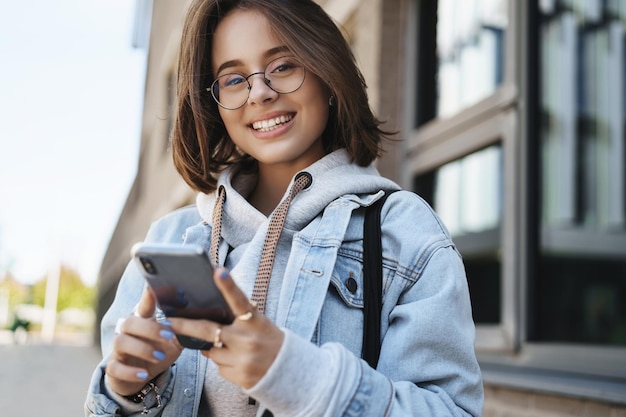 Ritratto del primo piano di attraente giovane donna caucasica in giacca di jeans in attesa della sua data utilizzare l'applicazione del telefono cellulare guardare la fotocamera con un sorriso raggiante in piedi sulla strada al mattino