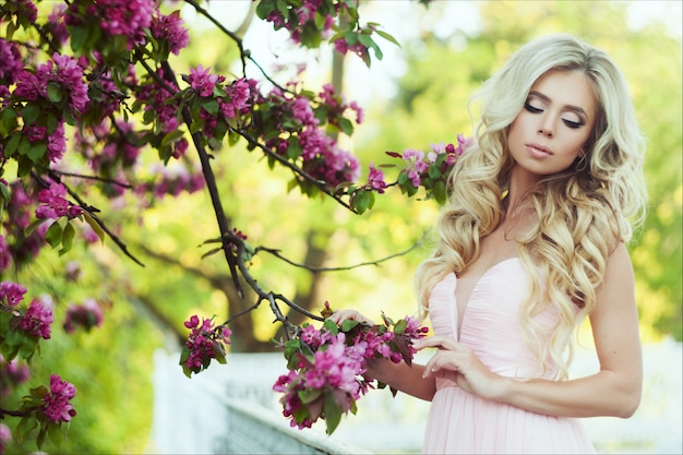 Ritratto del primo piano di bella donna con capelli biondi lunghi in un giardino floreale rosa di estate.