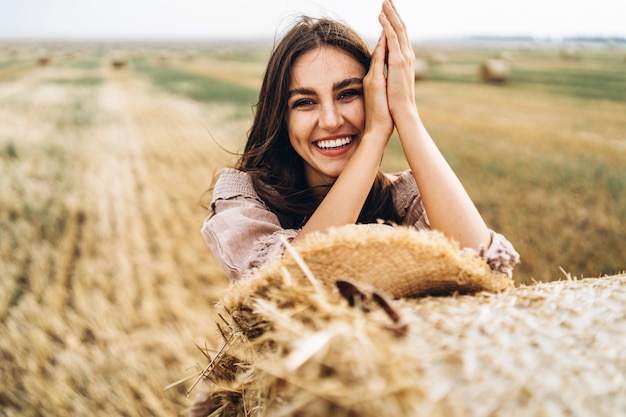 Ritratto del primo piano di bella donna sorridente con gli occhi chiusi. La bruna si appoggiò a una balla di fieno. Un campo di grano