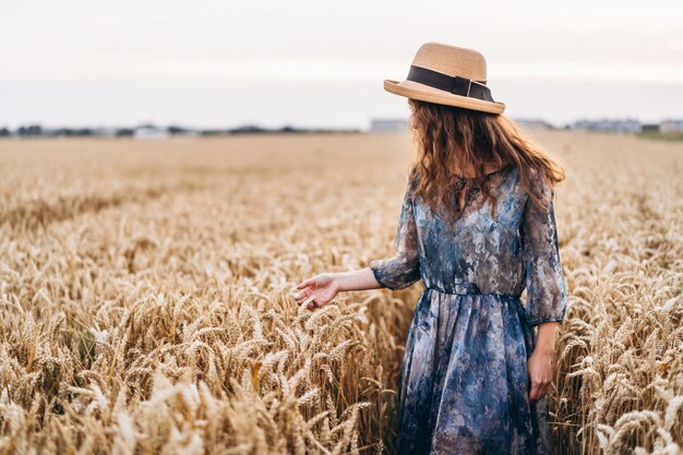 Ritratto del primo piano di bella giovane donna con capelli ricci. Donna in vestito e cappello che stanno nel campo di frumento